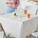 A woman pouring orange liquid from a bottle into a glass on a table with a white tissue/poly table cover.