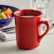 A close-up of a Tuxton Concentrix cayenne mug with liquid in it next to a bowl of fruit.