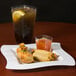 A Fineline white plastic square plate with a glass of iced tea and a glass of orange liquid on it, with food on the plate.