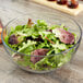 An Anchor Hocking glass bowl filled with a salad of leafy greens on a wood table.