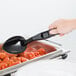 A person holding a black Cambro salad bar spoon over a tray of food.