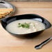 A black Fineline plastic bowl filled with soup, bread sticks, and a silver spoon.