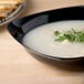 A black Fineline disposable plastic bowl filled with soup and topped with parsley.