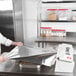 A man in a white shirt uses a Tablecraft KenKut film and foil dispenser to wrap a metal tray of food.