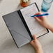 A person holding a Tablecraft black vinyl server book and writing in a checkbook.