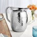 An American Metalcraft hammered stainless steel water pitcher on a table with a white tablecloth.