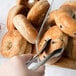 A person using tongs to grab a bagel from a Cal-Mil food bin.