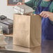 A man in a blue apron opening a Duro brown paper shopping bag to put a white styrofoam container inside.