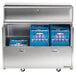 A Traulsen stainless steel milk cooler in a school kitchen with blue bins inside.