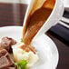 An American Metalcraft white porcelain gravy boat pouring gravy over a plate of meat.