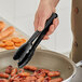 A person holding Mercer Culinary black high temperature plastic tongs over food.