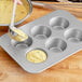 A person using a silver bowl to pour batter into a Chicago Metallic Texas Jumbo Muffin Pan.