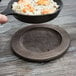 A hand holding a Lodge round wood underliner with a pan of rice and shrimp.