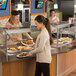 A woman serving food in a hospital cafeteria using a Hatco Glo-Ray heated stone warmer shelf.