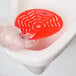 A gloved hand using a red Lavex Cherry Scent Deodorized Gel Urinal Screen brush in a urinal.