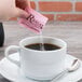 A hand pouring a Regal Pink Saccharin packet into a white cup of liquid.