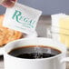 A hand holding a Regal cane sugar packet over a cup of coffee with brown liquid and sugar.