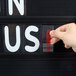 A person using a hand to change a sign on an Aarco Rocker two-sided black letterboard.