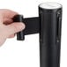A person holding a black Aarco crowd control stanchion with a black retractable belt.