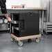 A man using a Cambro coffee beige Camdolly to transport a black container of food.