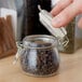 A hand holding an American Metalcraft glass miniature apothecary jar filled with brown cloves.