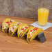 A row of American Metalcraft stainless steel taco holders with tacos on a table next to a glass of orange juice.