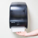 A person's arm and hand using a San Jamar black hands-free roll towel dispenser.