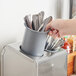 A hand holding a Steril-Sil gray plastic flatware cylinder filled with spoons.