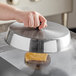 A hand holding an American Metalcraft stainless steel basting cover over a cheeseburger on a grill.