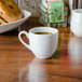 A CAC white bone china cup filled with brown liquid on a table with a bagel.