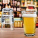 A close-up of an Anchor Hocking Clarisse stackable cooler glass full of beer with foam on a table next to a stack of glasses.