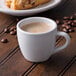A white espresso cup filled with brown liquid on a saucer with a cup of coffee beans on a wooden table.
