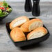 A black Milano square bowl filled with bread rolls on a table.