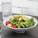 A Milano white melamine bowl filled with salad next to a glass of water.