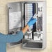 A woman in blue gloves is opening a metal cabinet holding a Bobrick sanitary napkin and tampon vendor.