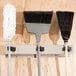 A Bobrick stainless steel mop and broom rack holding three brooms on a wall.