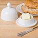 A CAC porcelain butter dish with a lid next to a bowl of butter and a bagel on a wooden table.