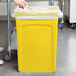 A hand putting a plastic bag in a yellow Rubbermaid Slim Jim trash can.