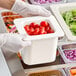 A person in gloves holding a white Cambro food pan filled with tomatoes.