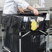 A person in a white coat and gloves rolling a black Rubbermaid cart with a black cover.