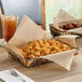 A basket of food on a table with a glass of brown liquid and a fork on a napkin, lined with a natural kraft deli wrap.