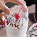 A clear plastic dome lid with a 2-inch hole being placed on a cup topped with whipped cream, chocolate drizzle, and a cherry.
