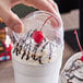 A clear PET dome lid with a 2-inch hole being placed on a cup topped with whipped cream, chocolate drizzle, and a cherry.