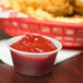 A plastic Solo translucent polystyrene portion cup filled with ketchup next to a basket of french fries.