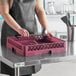 A woman in an apron holding a Vollrath Traex burgundy flatware rack over a dishwasher.