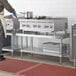 A man in a white apron standing on a Regency stainless steel equipment stand in a professional kitchen.