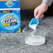 A hand using a blue OxiClean measuring spoon to pour white powder into a container of liquid.