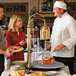 A chef in a white coat using a Hatco carving station to serve meat to a woman in a red blouse.