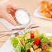 A person pouring dressing on a bowl of salad using a Carlisle stainless steel sauce cup.