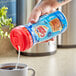 A hand pouring white powder from a blue plastic bottle into a cup of coffee.